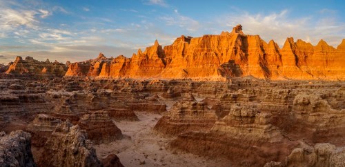 A panoramic view of rugged, orange-hued rock formations under a blue sky at sunset in a desert landscape.