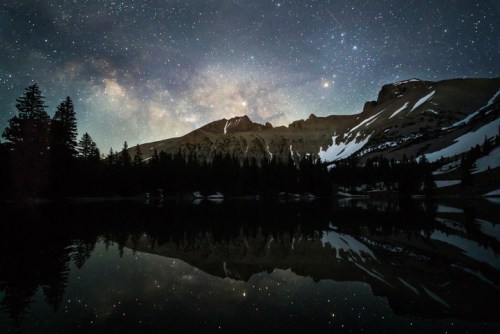 A serene night scene of mountains and trees reflected in a calm lake, under a starry sky with the Milky Way visible.