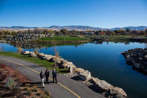 Three people walk along a path by a serene lake, surrounded by rocks and mountains under a clear blue sky.