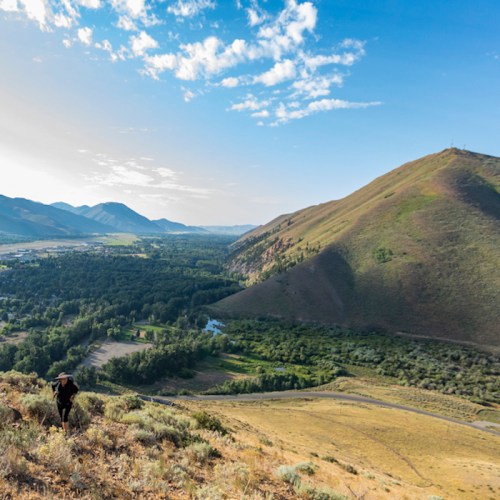 A person hiking up a grassy hill, overlooking a valley with mountains and a river in the distance under a blue sky.