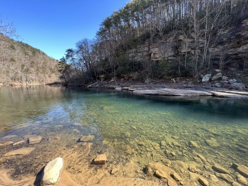 A serene river scene with clear water, rocky shore, and wooded cliffs under a blue sky.