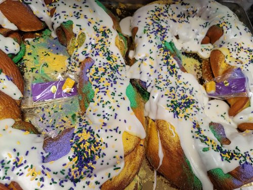 A close-up of a colorful King Cake topped with white icing and purple, green, and yellow sprinkles.