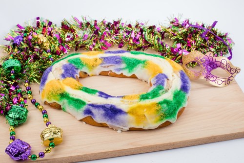 A colorful king cake with purple, green, and yellow icing, surrounded by festive Mardi Gras decorations and beads.