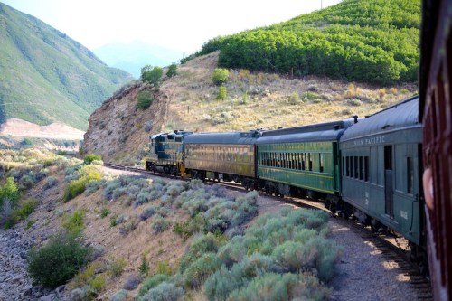 A vintage train winds through a mountainous landscape, surrounded by greenery and rocky terrain.