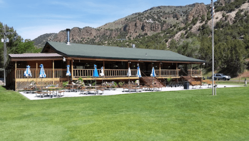 A rustic wooden restaurant with a green roof, surrounded by mountains and a grassy area, featuring outdoor seating.