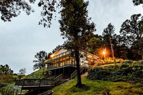 A modern house on a hillside, surrounded by trees, with warm lights glowing at dusk.