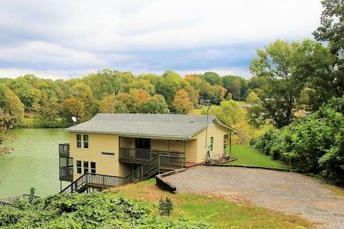 A yellow house by a lake, surrounded by trees with autumn foliage, under a cloudy sky.