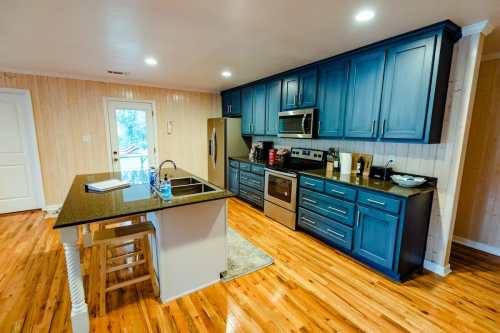 Modern kitchen with blue cabinets, stainless steel appliances, and a central island, featuring wooden floors and natural light.