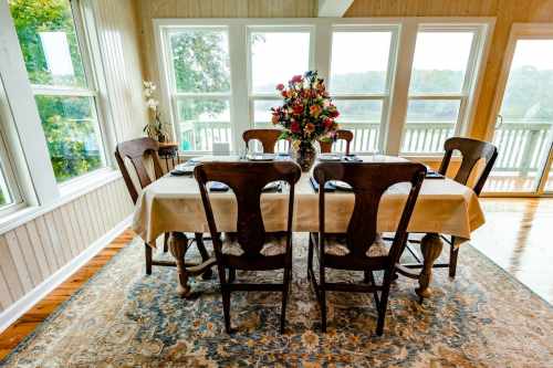 A dining table set with chairs and a floral centerpiece, surrounded by large windows with a view of nature.