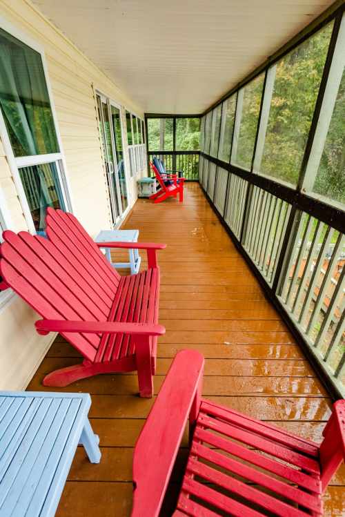 A screened porch with red and blue Adirondack chairs on a wooden floor, surrounded by greenery.
