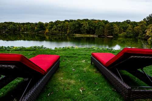 Two red lounge chairs sit on green grass overlooking a calm lake surrounded by trees under a cloudy sky.