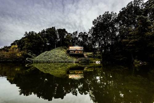 A serene house on a hill reflected in a calm lake, surrounded by lush trees and a cloudy sky.