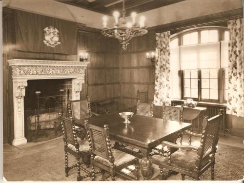 A vintage dining room with a wooden table, chairs, a fireplace, and large windows adorned with floral curtains.