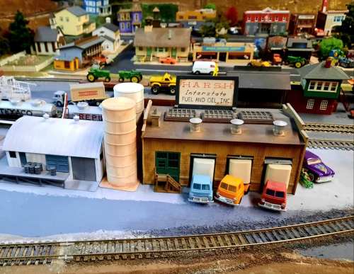 A detailed model of a fuel and oil facility with trucks, silos, and nearby buildings in a miniature train setup.