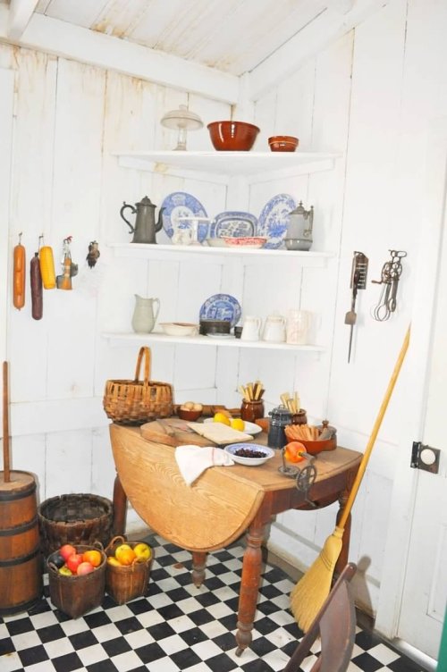 A rustic kitchen with a wooden table, shelves of dishes, baskets of fruit, and a broom on a checkered floor.