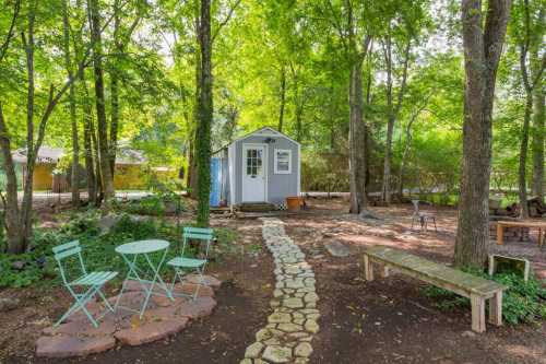 A cozy white shed surrounded by trees, with a stone path, green chairs, and a wooden bench in a serene outdoor setting.