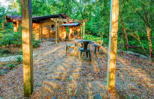 A cozy outdoor seating area with a table and chairs surrounded by trees and a rustic building in the background.