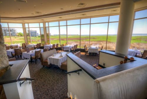 Elegant restaurant interior with white tablecloths, large windows showcasing a scenic view of greenery and water.
