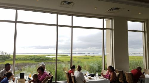 A restaurant interior with diners enjoying a view of a green landscape and cloudy sky through large windows.