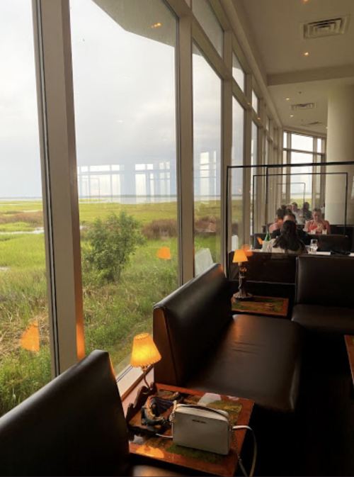 A cozy restaurant with large windows overlooking a green marsh, featuring dark seating and soft lighting.