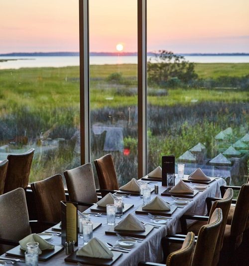 A dining table set for a meal, with a sunset view over a grassy landscape through large windows.