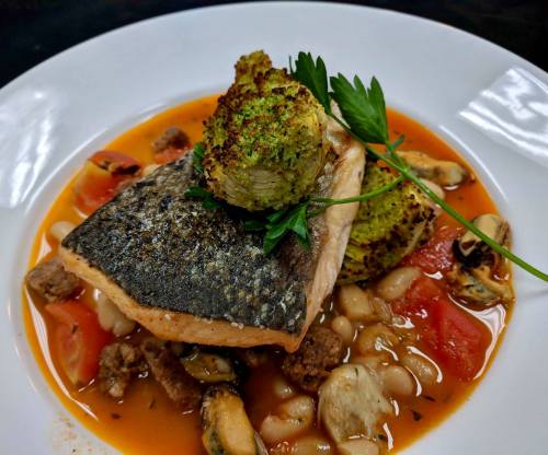 A plated dish featuring seared salmon, green herb crust, beans, tomatoes, and mussels in a savory broth.
