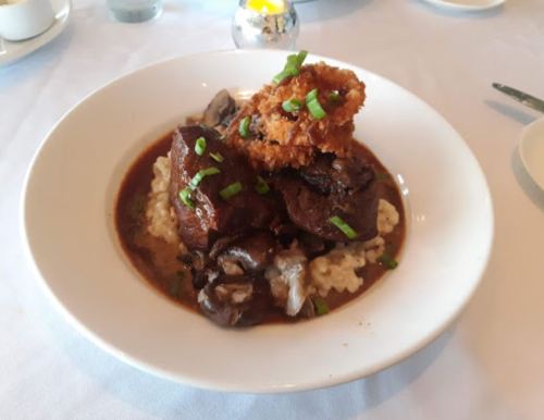 A plate of tender beef with mushrooms and risotto, topped with a crispy onion ring and garnished with green onions.