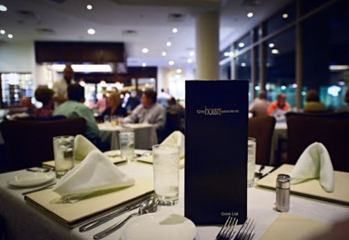 A restaurant table set with a menu, silverware, and glasses, with diners in the background. Soft lighting creates a cozy atmosphere.