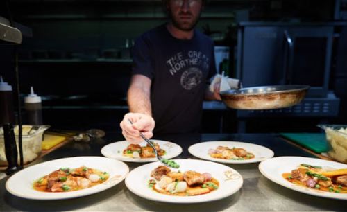 A chef plating dishes in a kitchen, focusing on a plate with meat and vegetables, surrounded by other prepared plates.
