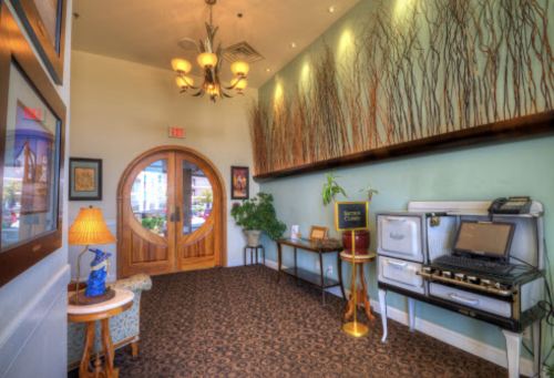A welcoming lobby with a round wooden door, decorative plants, and vintage furnishings, featuring warm lighting and wall art.
