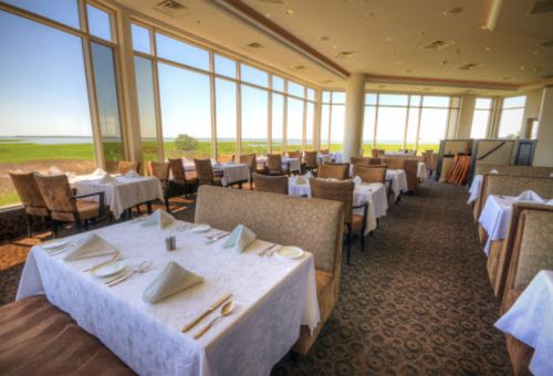 Elegant restaurant interior with white tablecloths, large windows, and a scenic view of green fields outside.