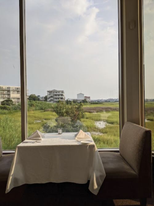 A cozy restaurant table set for two, overlooking a scenic view of green marshland and distant buildings.
