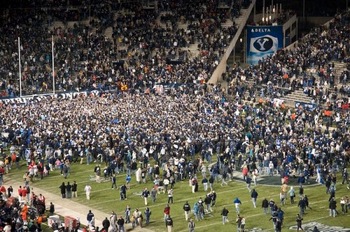 A large crowd of fans rushes the field in celebration after a football game, with stadium seating in the background.