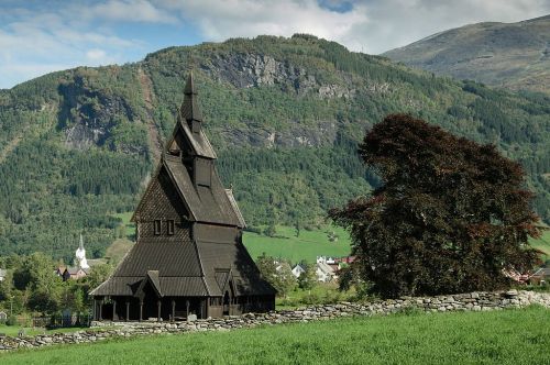 A wooden church with a tall spire, surrounded by lush green fields and mountains under a partly cloudy sky.