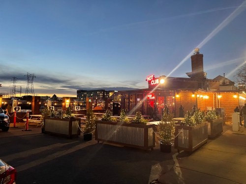 A lively restaurant with outdoor seating, illuminated by string lights, against a twilight sky.