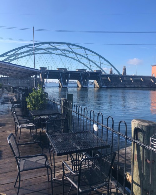 A riverside view featuring empty black metal tables and chairs, with a large bridge in the background under a clear blue sky.