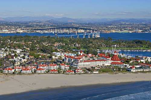 Aerial view of a coastal area featuring a hotel with red roofs, a bridge, and lush greenery along the shoreline.