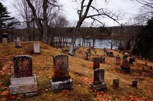 A serene cemetery with weathered gravestones on a hillside, overlooking a calm river and surrounded by bare trees.