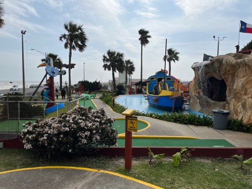 Colorful mini-golf course with palm trees, a pirate ship, and a water feature near the beach on a cloudy day.