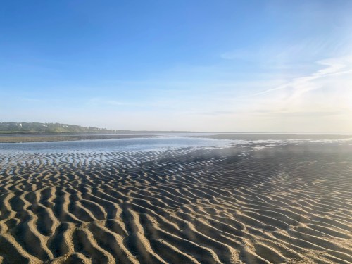 A serene beach scene with rippled sand and a calm, reflective water surface under a clear blue sky.