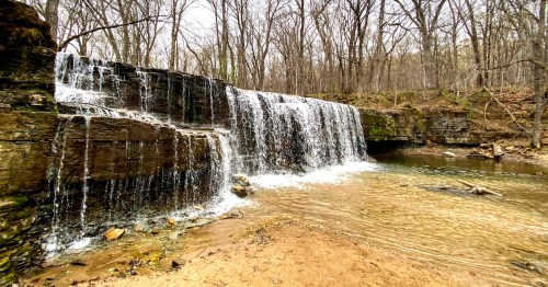 A serene waterfall cascading over rocky ledges into a clear stream, surrounded by bare trees in a tranquil forest setting.