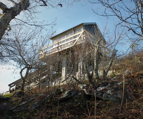 A wooden structure perched on stilts, surrounded by bare trees and rocky terrain under a clear blue sky.