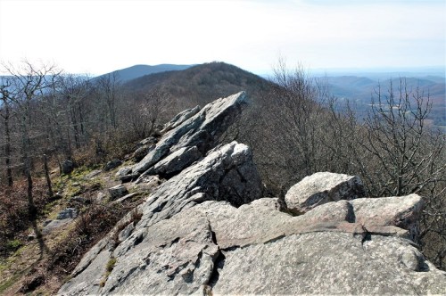 Rocky outcrop on a mountain ridge with bare trees and distant hills under a clear sky.