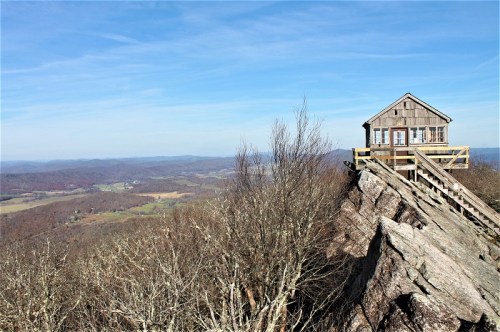 A wooden cabin on a rocky outcrop overlooks a vast valley and distant mountains under a clear blue sky.