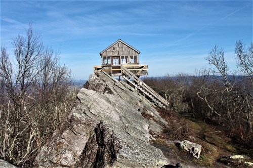 A wooden cabin perched on a rocky outcrop, surrounded by bare trees and a clear blue sky.