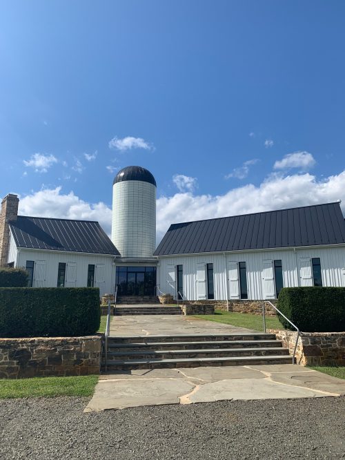 A modern white building with a cylindrical silo, surrounded by greenery and a stone pathway under a blue sky.