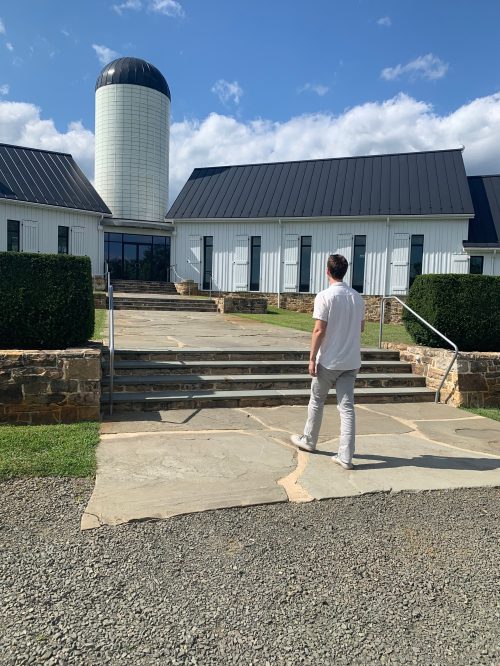A person walks up stone steps towards a modern barn with a silo under a blue sky.