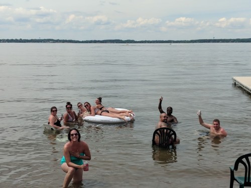 A group of people enjoying a sunny day in the water, some on a float, others wading, with a dock in the background.