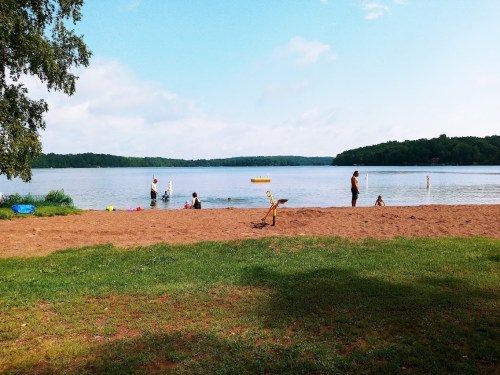 A sandy beach by a lake with people swimming and playing, surrounded by trees and a clear blue sky.