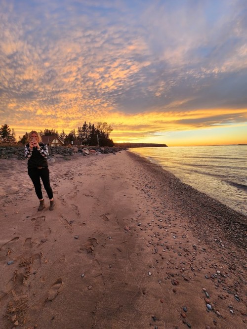 A person stands on a sandy beach at sunset, with colorful clouds and calm water in the background.